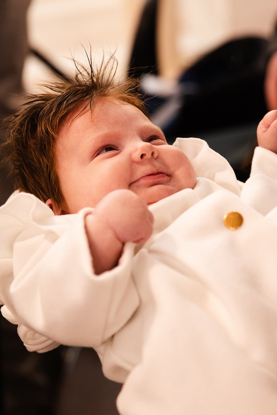 Uśmiechnięty noworodek w białym ubranku z pojedynczym złotym guzikiem, sfotografowany z dołu, z widocznymi włoskami na głowie.