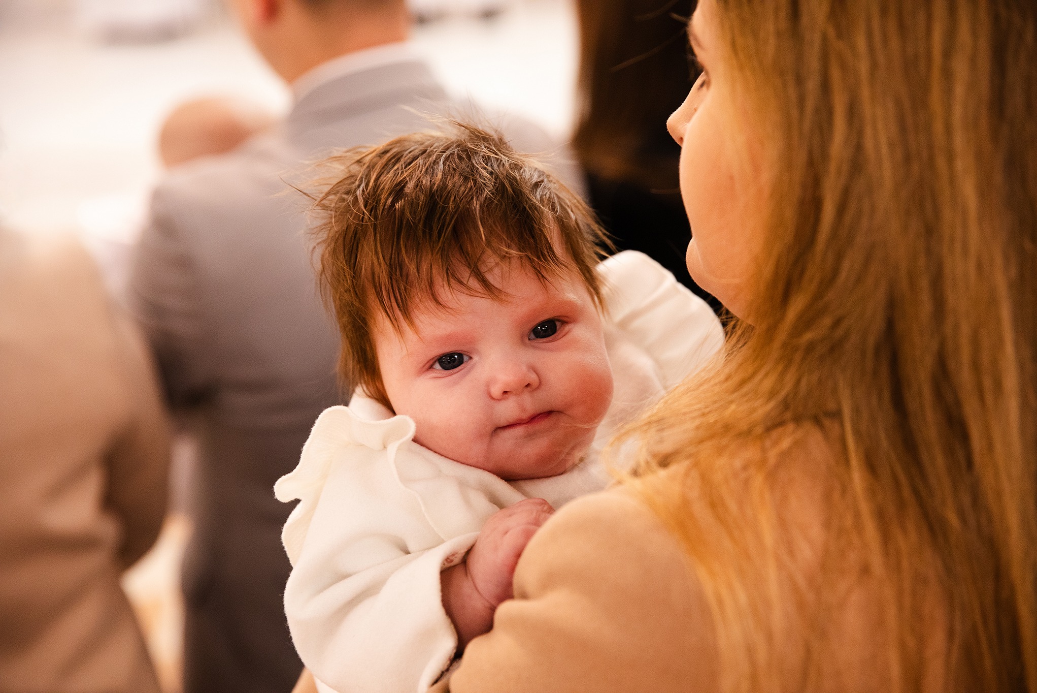 Portret niemowlęcia w białym ubranku, trzymanego przez osobę z długimi blond włosami. W tle rozmyte postacie, ciepłe oświetlenie.