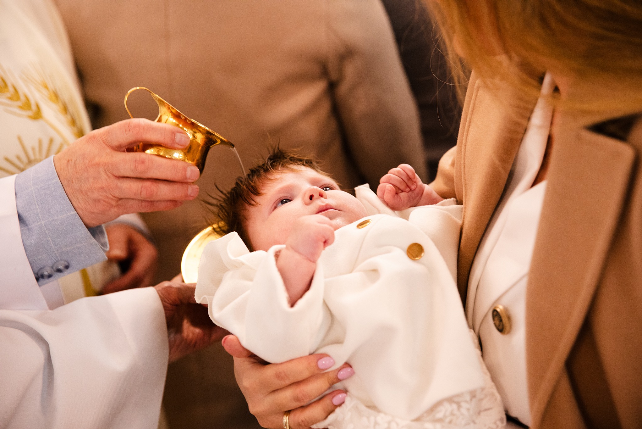 Chrzest dziecka: ksiądz polewa wodą główkę niemowlęcia ubranego w biały strój, trzymanego przez kobietę w beżowym płaszczu. Złoty dzbanek i jasne tło.