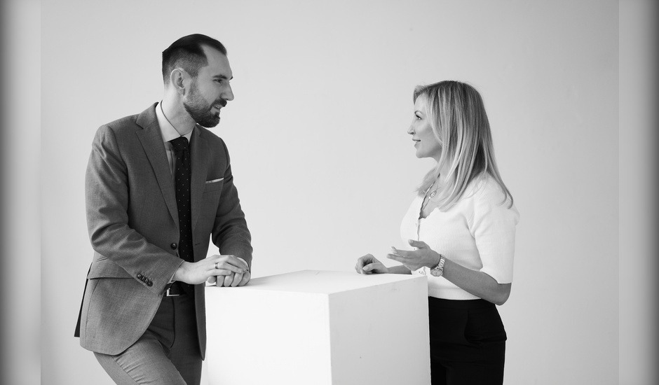 Czarno-białe zdjęcie przedstawia mężczyznę w garniturze i kobietę w bluzce, rozmawiających przy białym kubiku, prawdopodobnie w kancelarii.