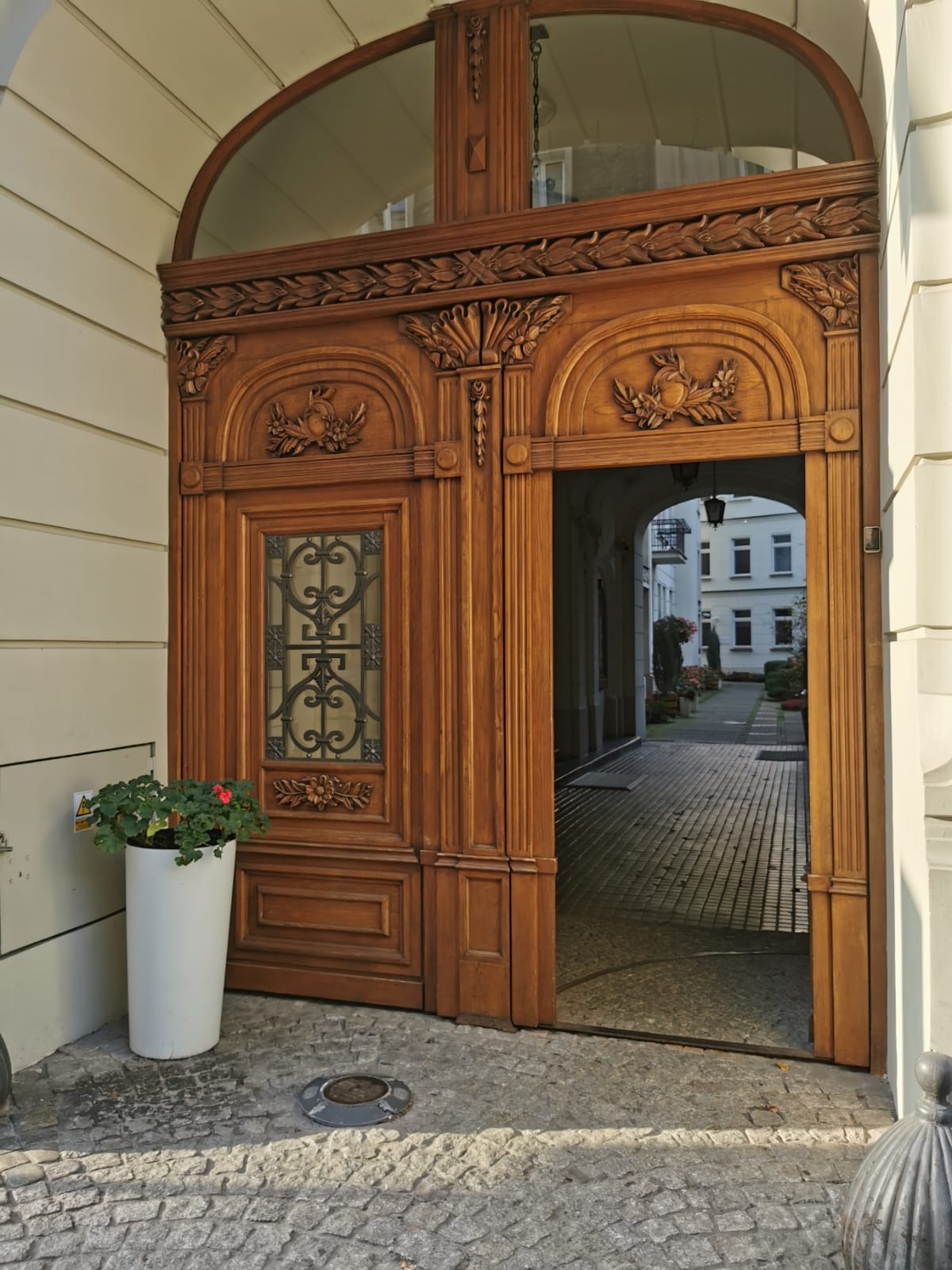 Otwarta, bogato zdobiona drewniana brama wejściowa z przeszkleniem w górnej części, widok na wewnętrzny dziedziniec z brukowaną ścieżką i budynkami w tle, donica z czerwonymi kwiatami po lewej...
