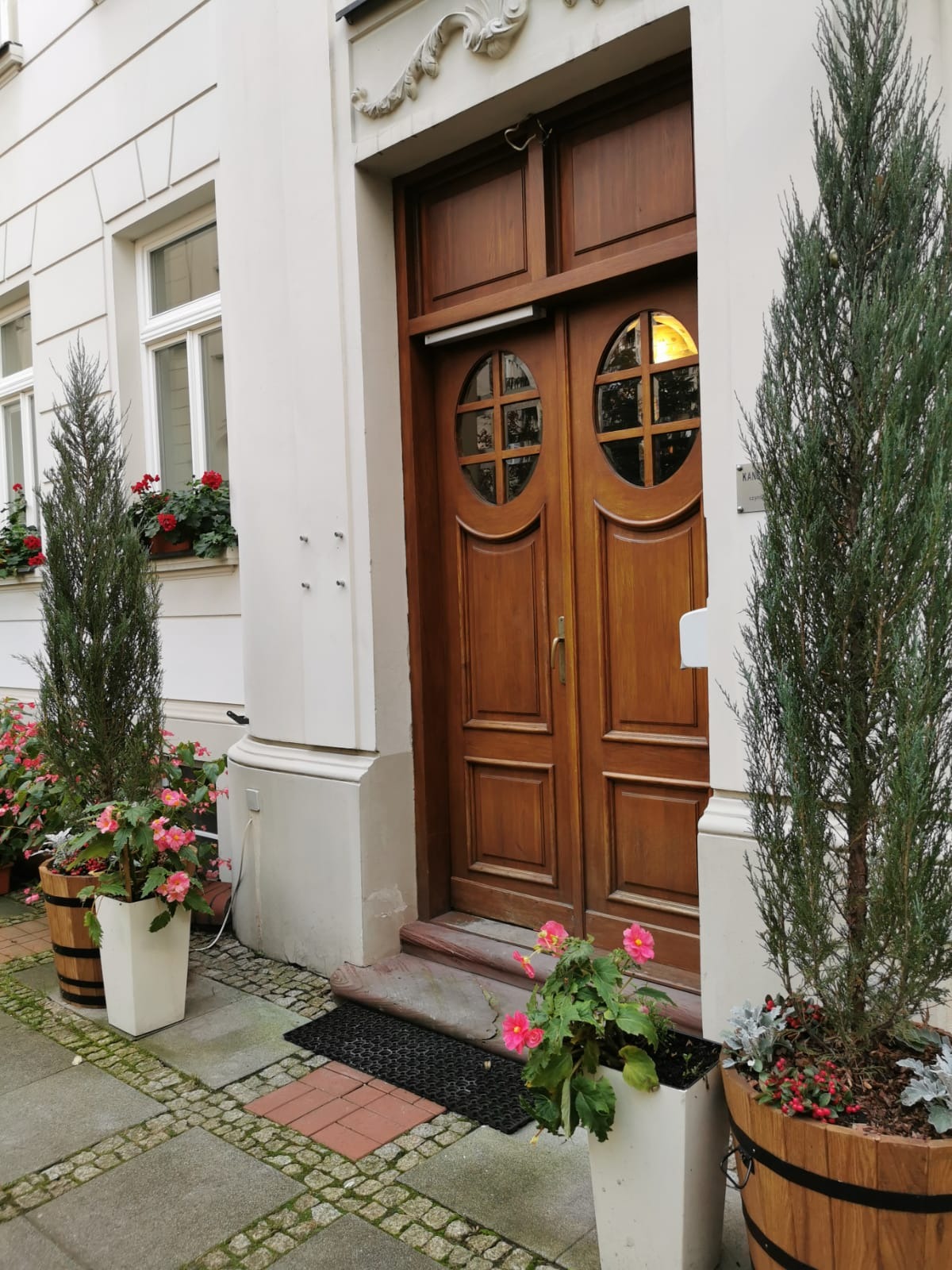 Wejście do budynku z drewnianymi drzwiami z okrągłymi okienkami, ozdobione roślinami w donicach, z widoczną tabliczką informacyjną po prawej stronie.