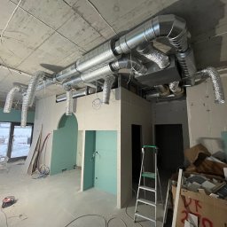 Lesław Prabucki - Wnętrze w trakcie prac wykończeniowych: widoczne instalacje wentylacyjne pod sufitem, ściany z płyt karton-gips, drabina i materiały budowlane. Surowy stan lokalu.