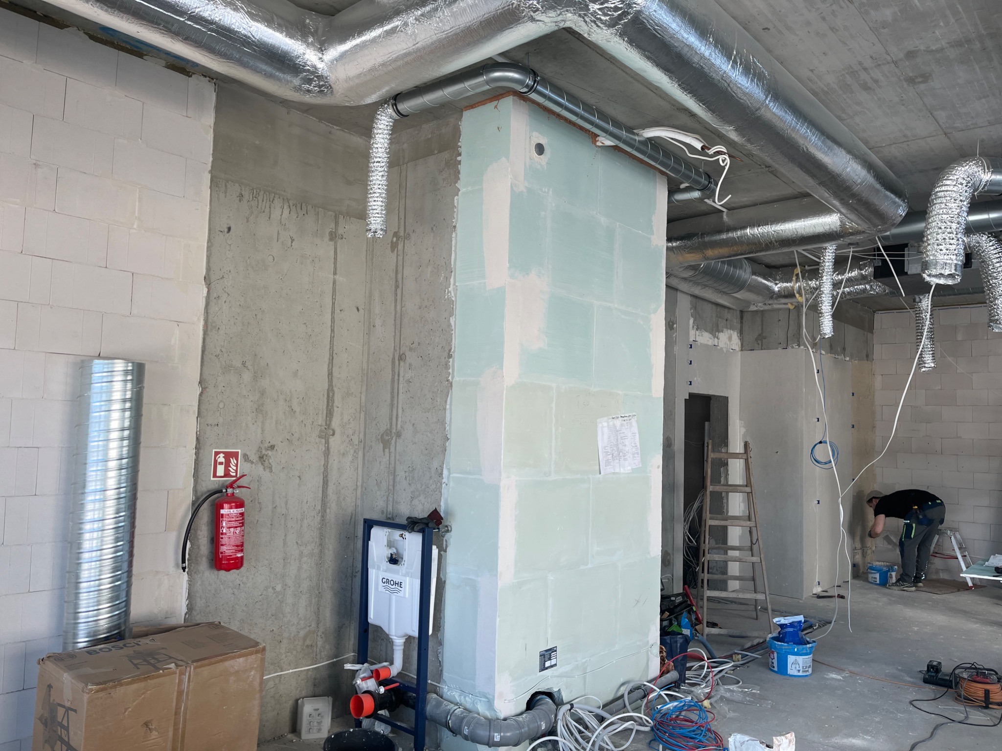 Wnętrze w trakcie prac wykończeniowych: widoczne instalacje wentylacyjne, obudowa z płyt G-K, instalacja sanitarna, ściany z betonu komórkowego i pracownik w tle.