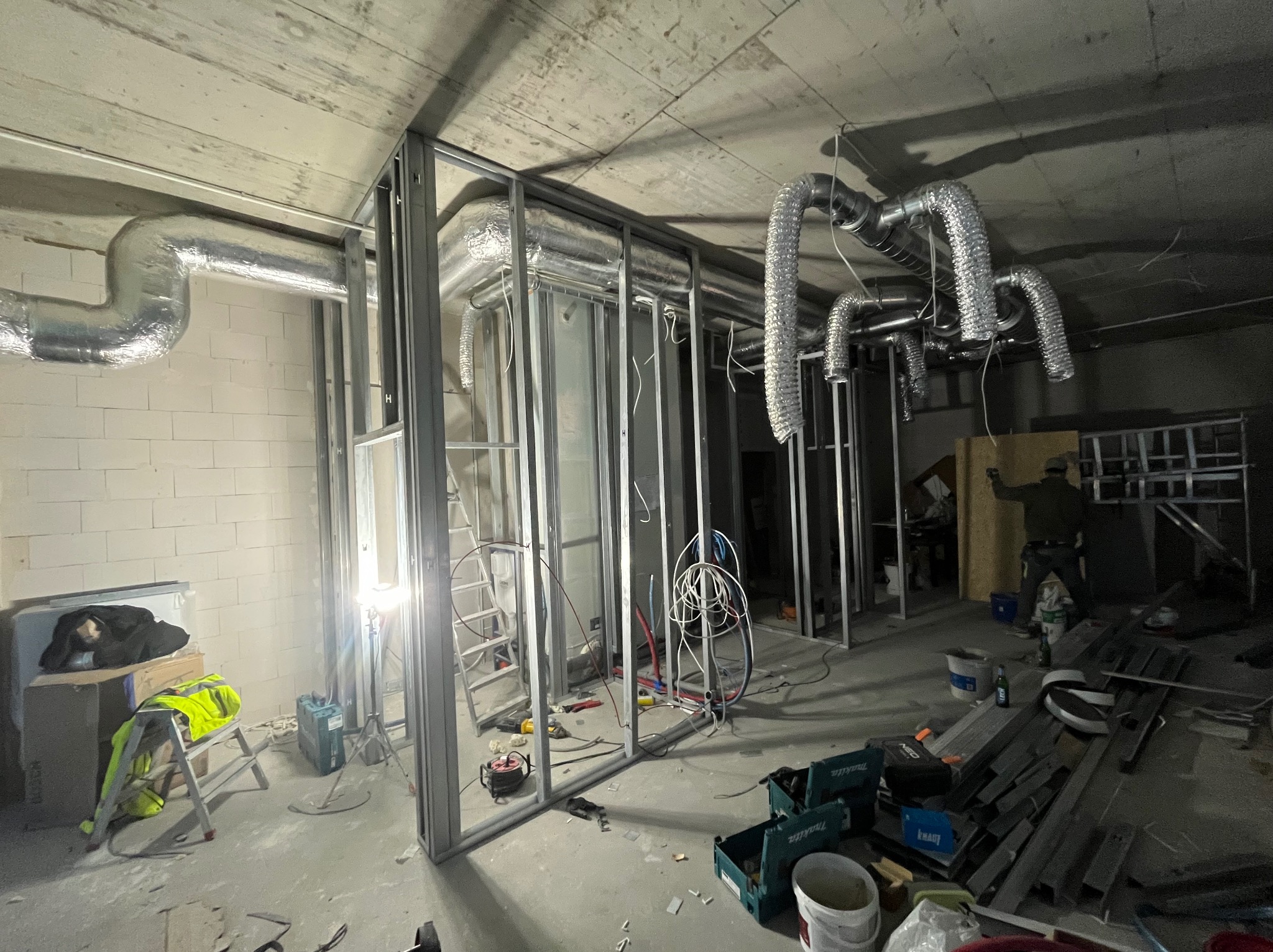 Wnętrze w trakcie prac: metalowe profile ścian działowych, instalacja wentylacyjna pod sufitem, drabina i narzędzia. Widoczne elementy stanu surowego.