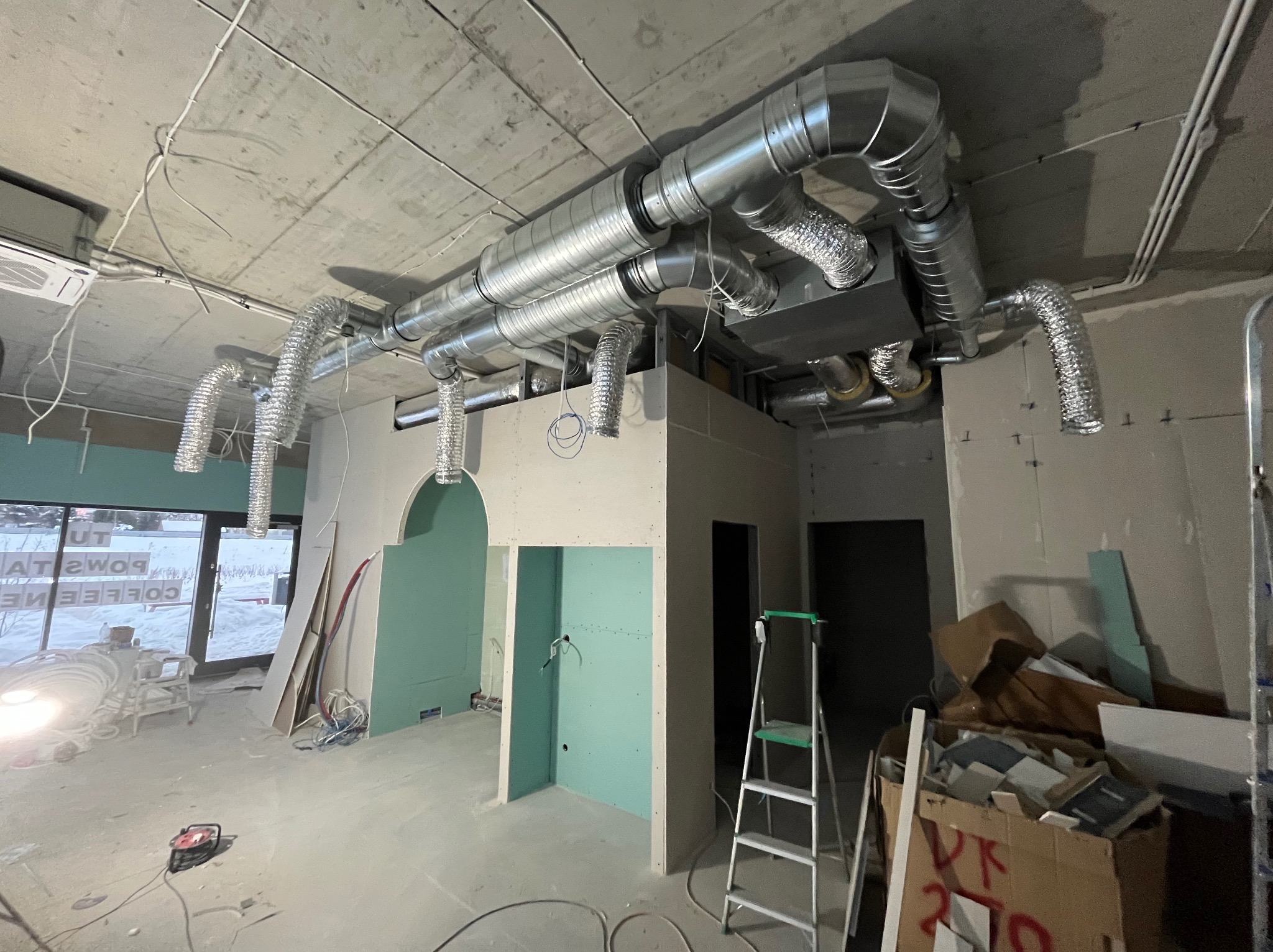 Wnętrze w trakcie prac wykończeniowych: widoczne instalacje wentylacyjne pod sufitem, ściany z płyt karton-gips, drabina i materiały budowlane. Surowy stan lokalu.