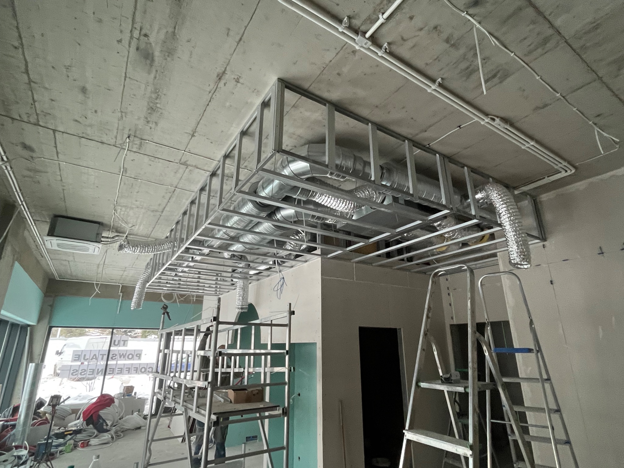 Metalowa konstrukcja sufitu podwieszanego z widocznymi przewodami wentylacyjnymi, drabiny i częściowo zamontowane płyty gipsowo-kartonowe w surowym wnętrzu.