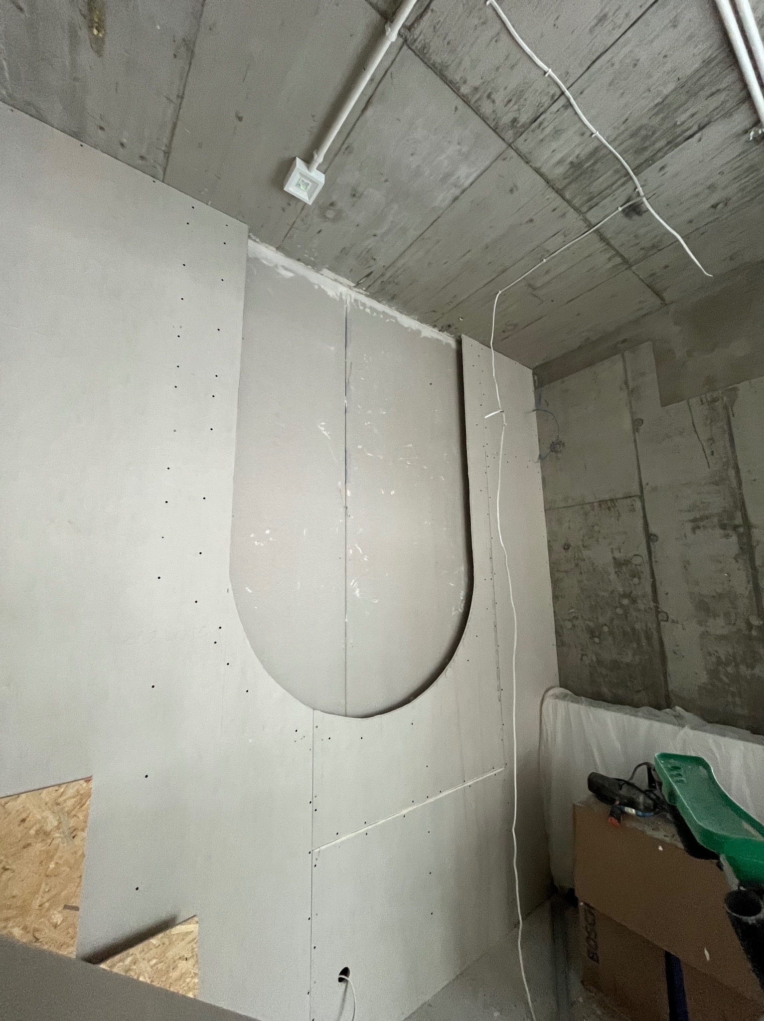 Montaż płyty gipsowej z wycięciem w kształcie łuku na ścianie w surowym wnętrzu. Widoczne narzędzia i instalacje elektryczne. Praca w toku.