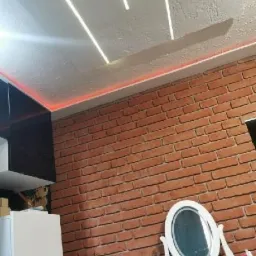 Sufit z oświetleniem LED wzdłuż krawędzi i liniowymi lampami, ściana z cegły, toaletka z owalnym lustrem.