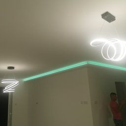 Pro-rem uslugi remontowe - Nowoczesne oświetlenie wnętrza: Dwa wiszące, białe, spiralne żyrandole LED oraz zielone oświetlenie LED podsufitowe, widoczny fragment ściany i sufitu, oraz osoba w tle.