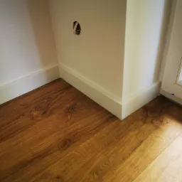 Fragment rogu pokoju z drewnianą podłogą, białą listwą przypodłogową i otworem na kabel w ścianie