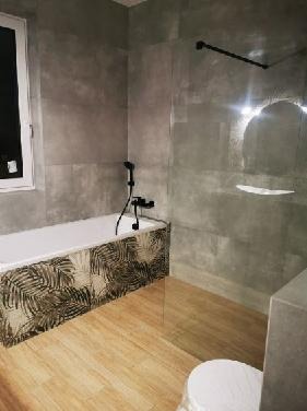 Nowoczesna łazienka z wanną obudowaną płytkami z motywem liści, szklaną ścianką prysznicową i drewnopodobną podłogą; minimalistyczna armatura w kolorze czarnym.