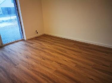 Pusty pokój po remoncie z nową podłogą z paneli w odcieniach brązu, jasne ściany, widok na zewnątrz przez okno balkonowe