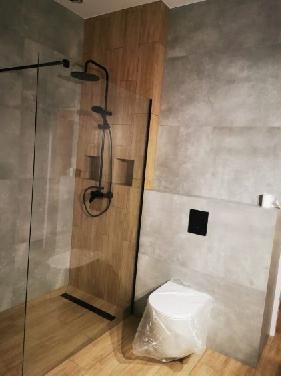 Nowoczesna łazienka z kabiną prysznicową typu walk-in, czarną armaturą i drewnopodobnymi płytkami na ścianie, toaleta w folii ochronnej.
