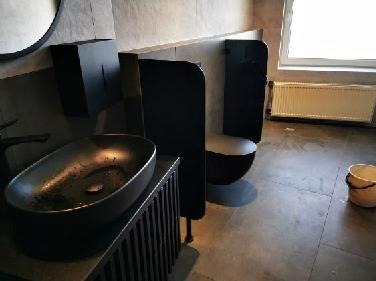 Nowoczesna łazienka w odcieniach szarości i czerni z owalną umywalką na szafce z lamelami, wiszącą toaletą za czarną przegrodą, grzejnikiem pod oknem i wiaderkiem na podłodze