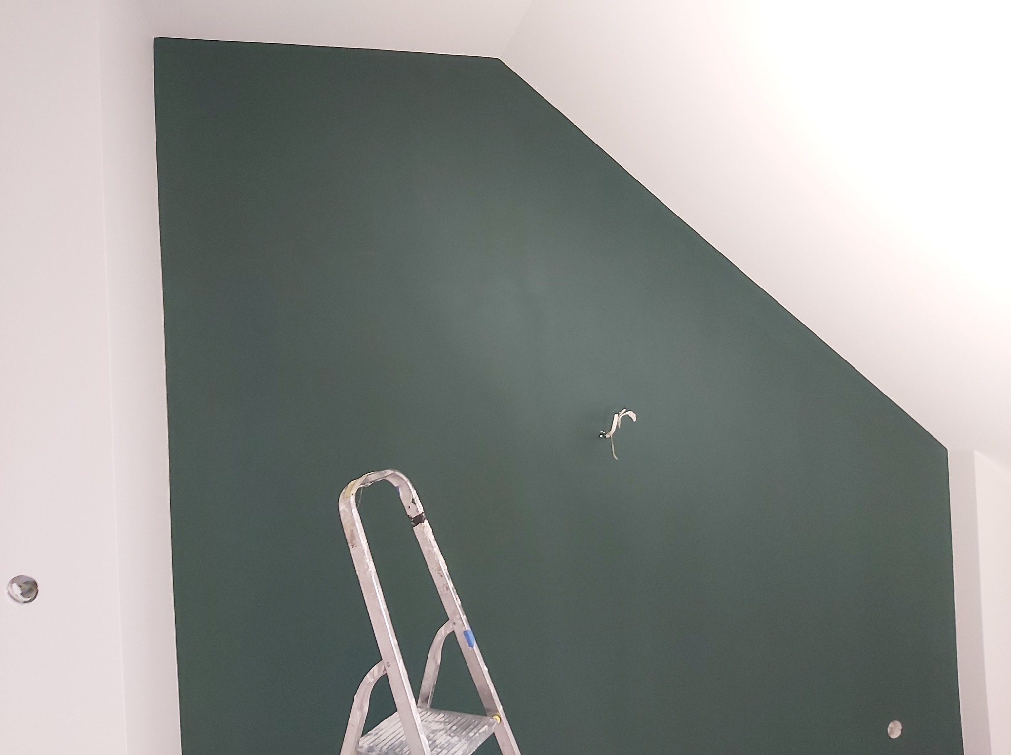 Świeżo pomalowana, ciemnozielona ściana ze skosem, z widoczną drabinką malarską i wystającym kablem elektrycznym.