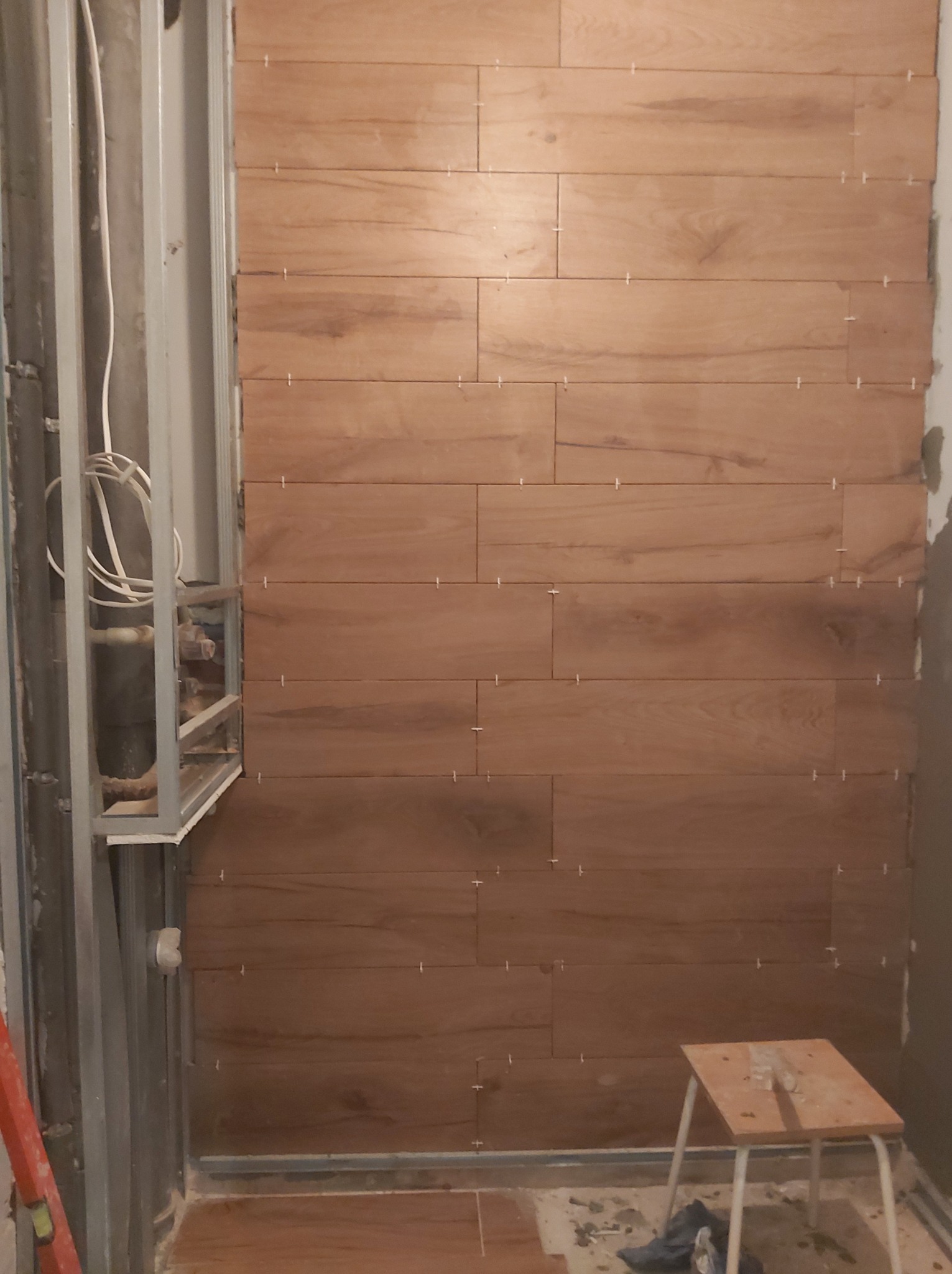 Układanie płytek imitujących drewno na ścianie w łazience, widoczny metalowy stelaż z instalacją hydrauliczną i mały stołek roboczy