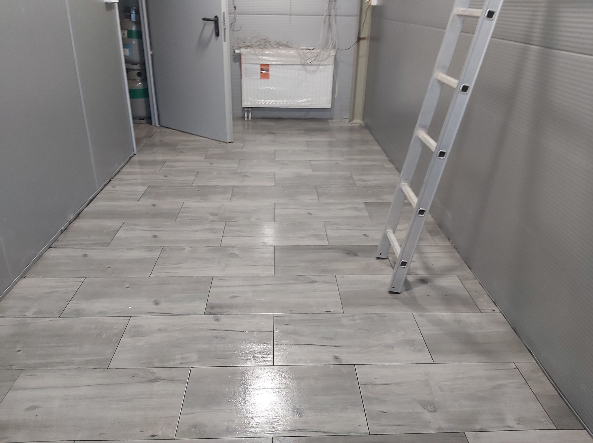 Wykończone wnętrze z szarymi płytkami imitującymi drewno na podłodze, drabina oparta o ścianę, widoczny grzejnik i instalacje w tle.