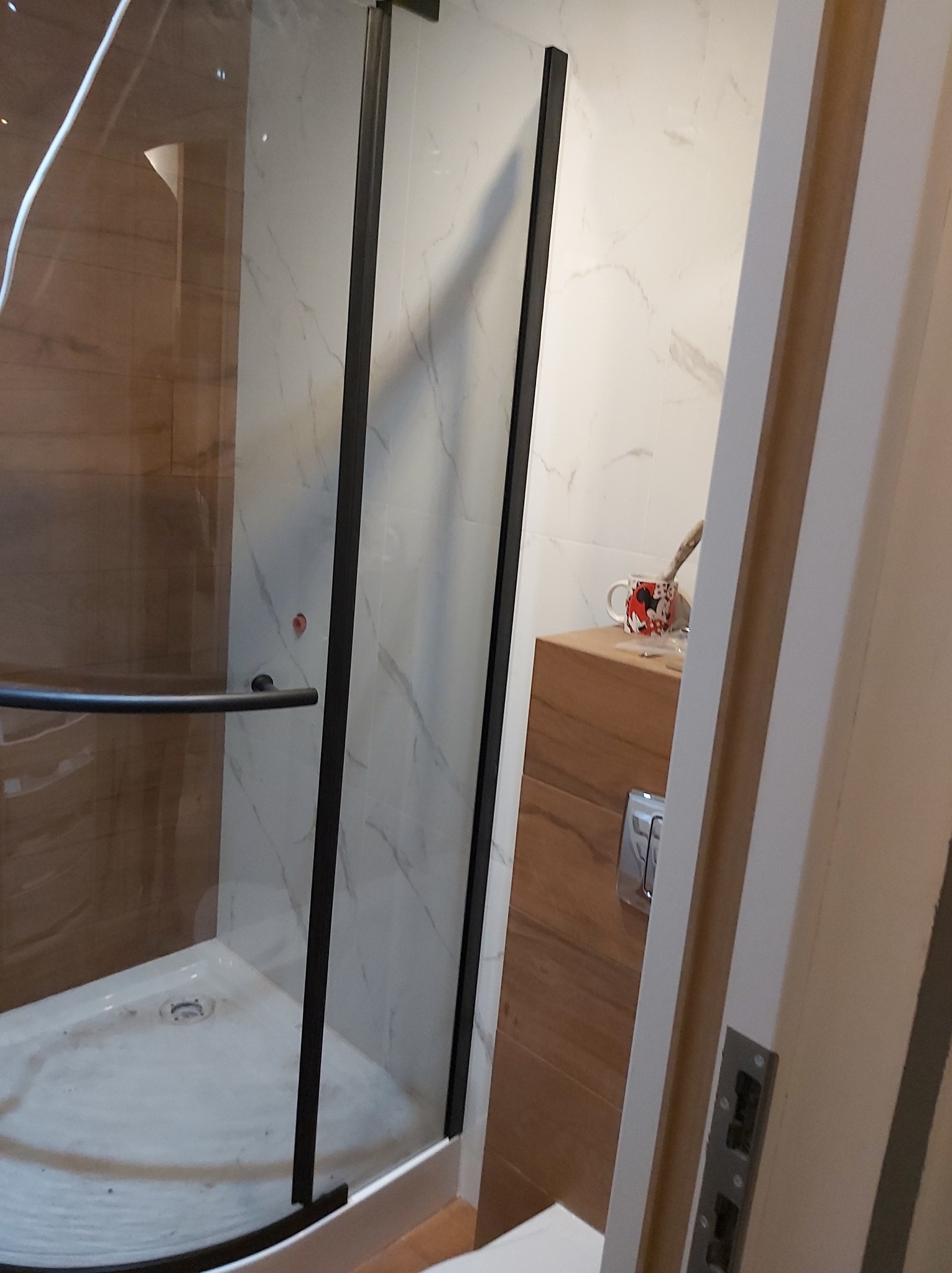 Narożna kabina prysznicowa z czarnymi profilami, białe płytki imitujące marmur na ścianach, drewniana szafka z umywalką i czerwony kubek z motywem Myszki Miki na blacie, widok zza otwartych drzwi.
