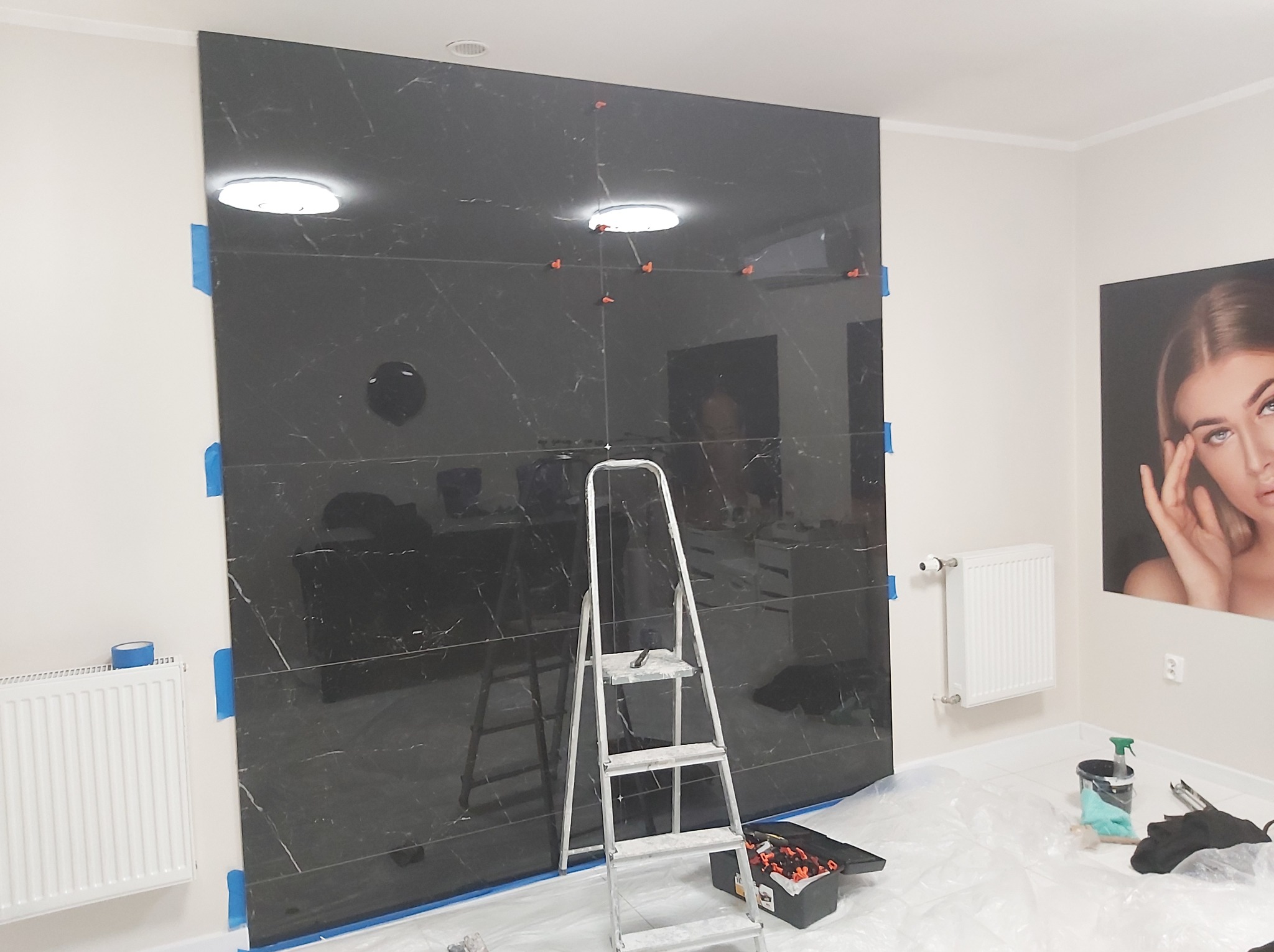 Montaż czarnych płytek imitujących marmur na ścianie w łazience, widoczne narzędzia glazurnicze, drabina i zabezpieczenia podłogi folią.