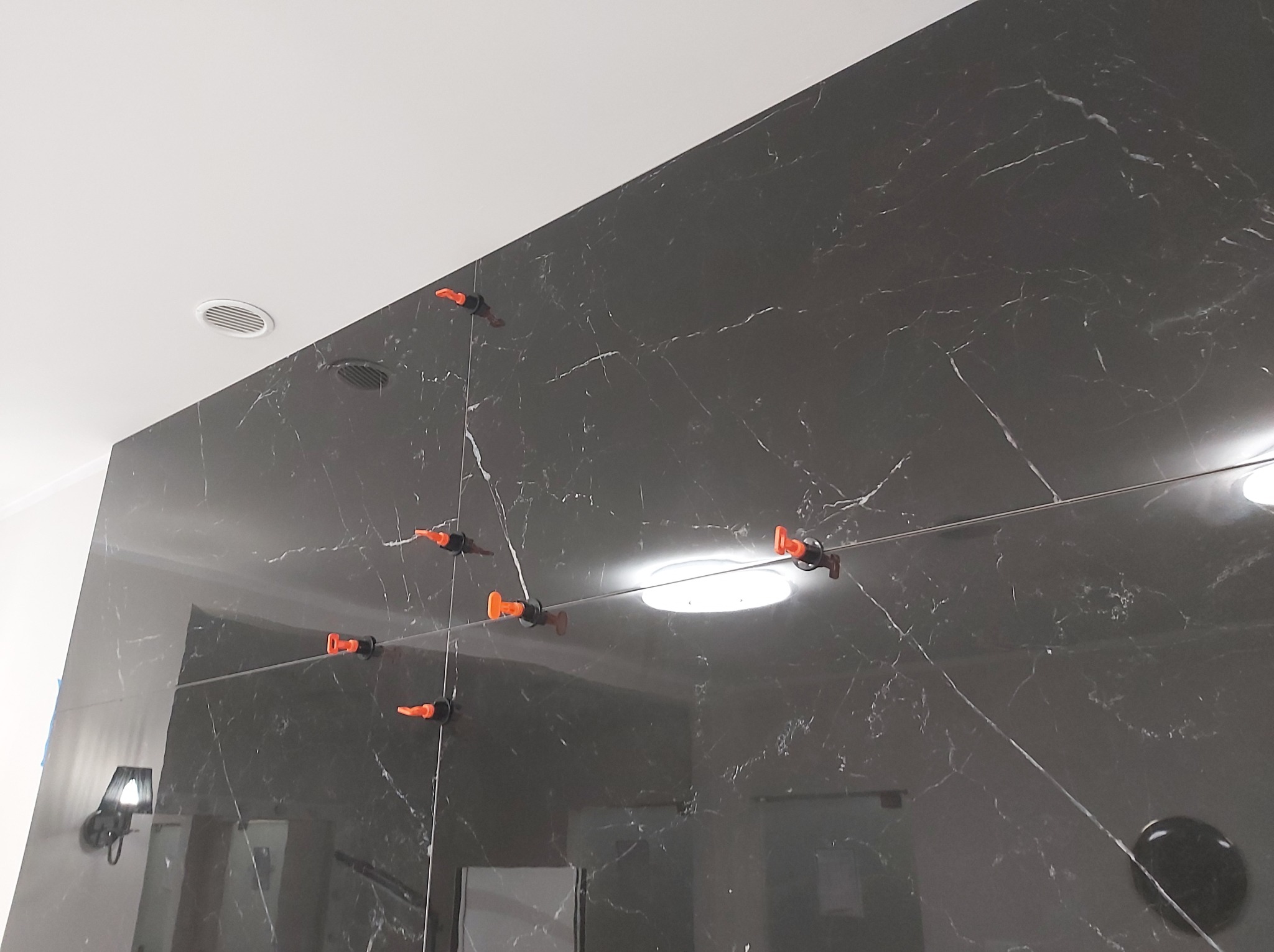 Montaż wielkoformatowych, ciemnych płytek imitujących marmur na ścianie, z użyciem systemu poziomowania płytek z pomarańczowymi klinami i metalowymi listwami.