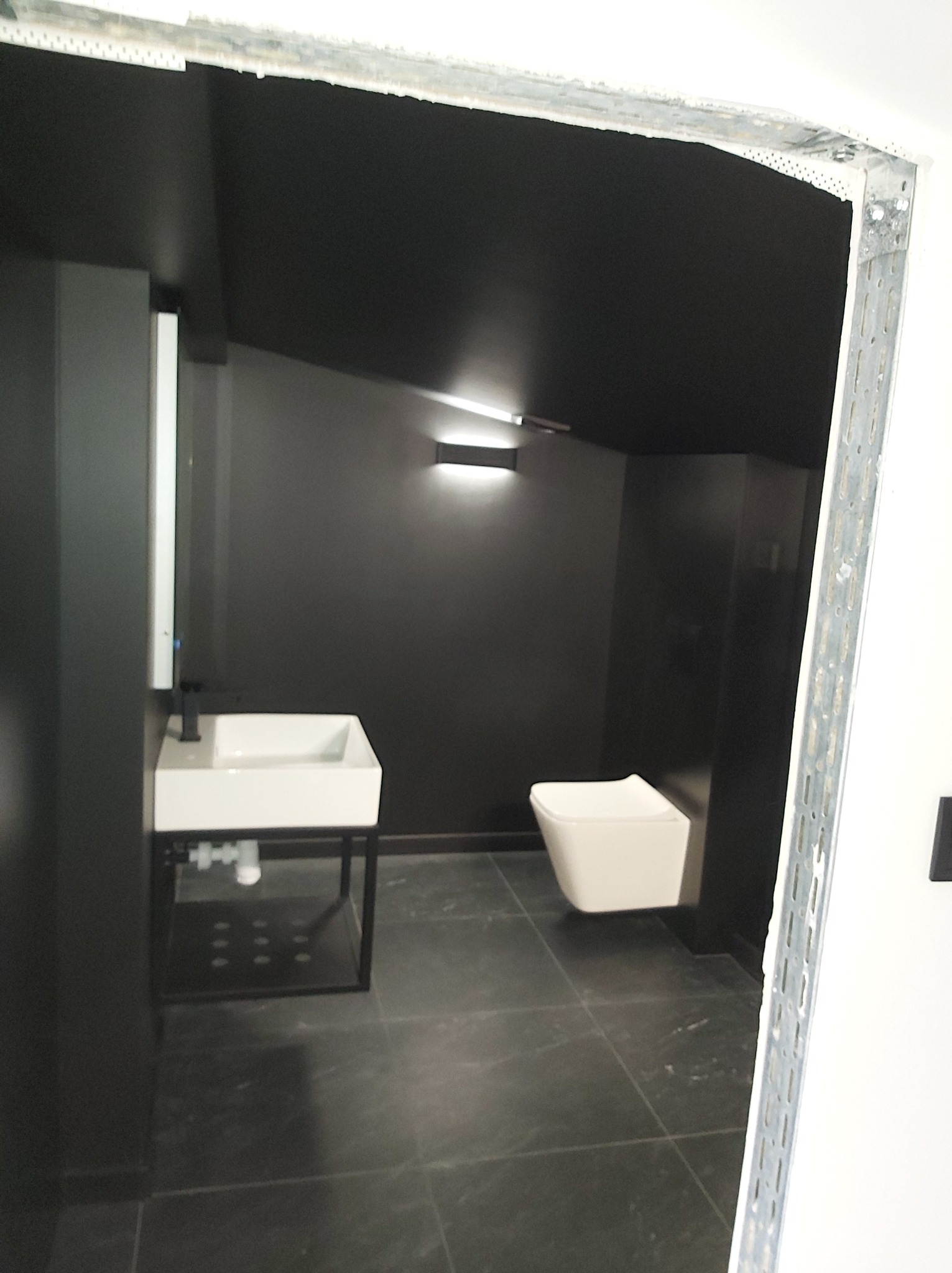 Nowoczesna łazienka w odcieniach szarości z wiszącą toaletą, umywalką na metalowym stelażu i oświetleniem punktowym, widoczna przez metalową ramę drzwi w trakcie montażu.