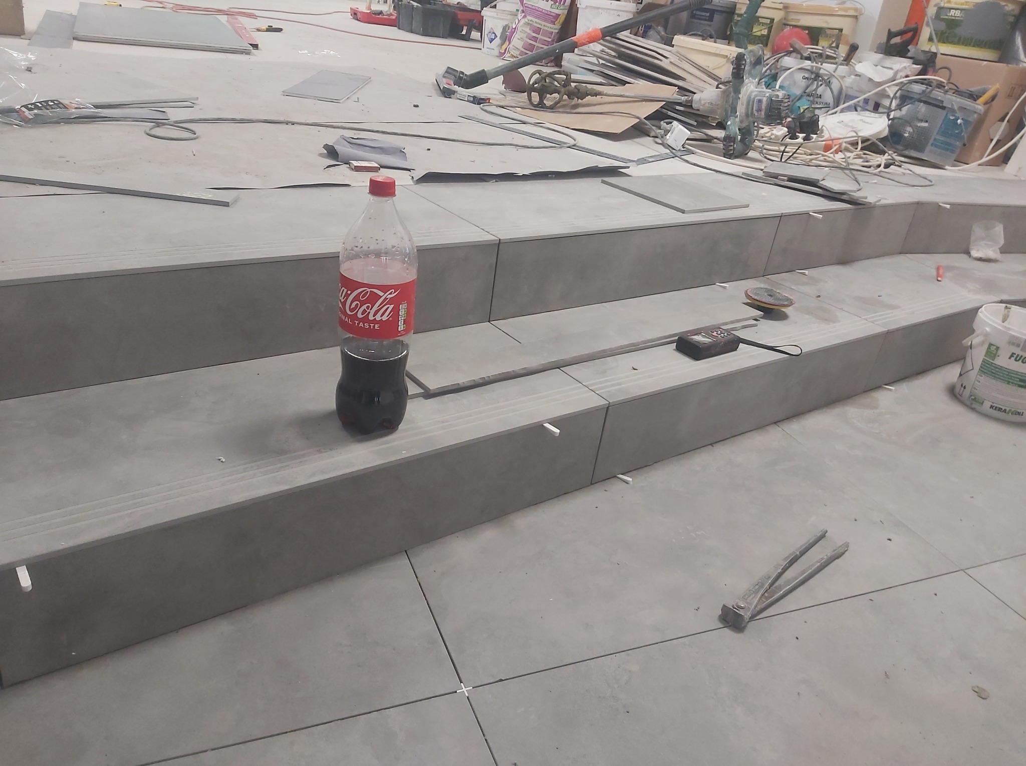 Montaż płytek na schodach w trakcie prac, widoczna butelka Coca-Coli, poziomica, narzędzia glazurnicze i zaplecze budowy z narzędziami.