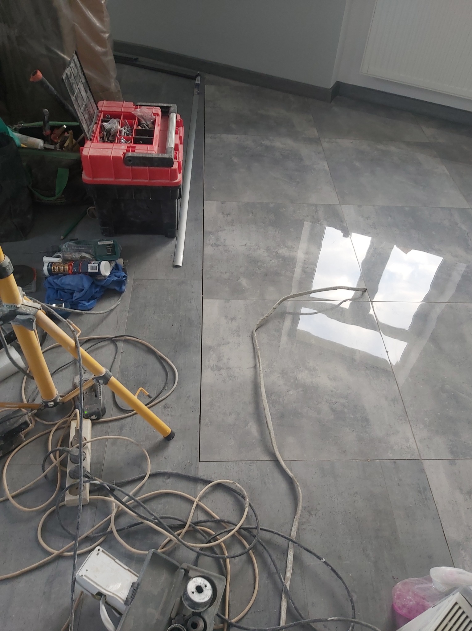 Szare płytki podłogowe z połyskiem, odbijające okno, z widocznymi narzędziami pracy, skrzynką narzędziową i kablem elektrycznym na podłodze.