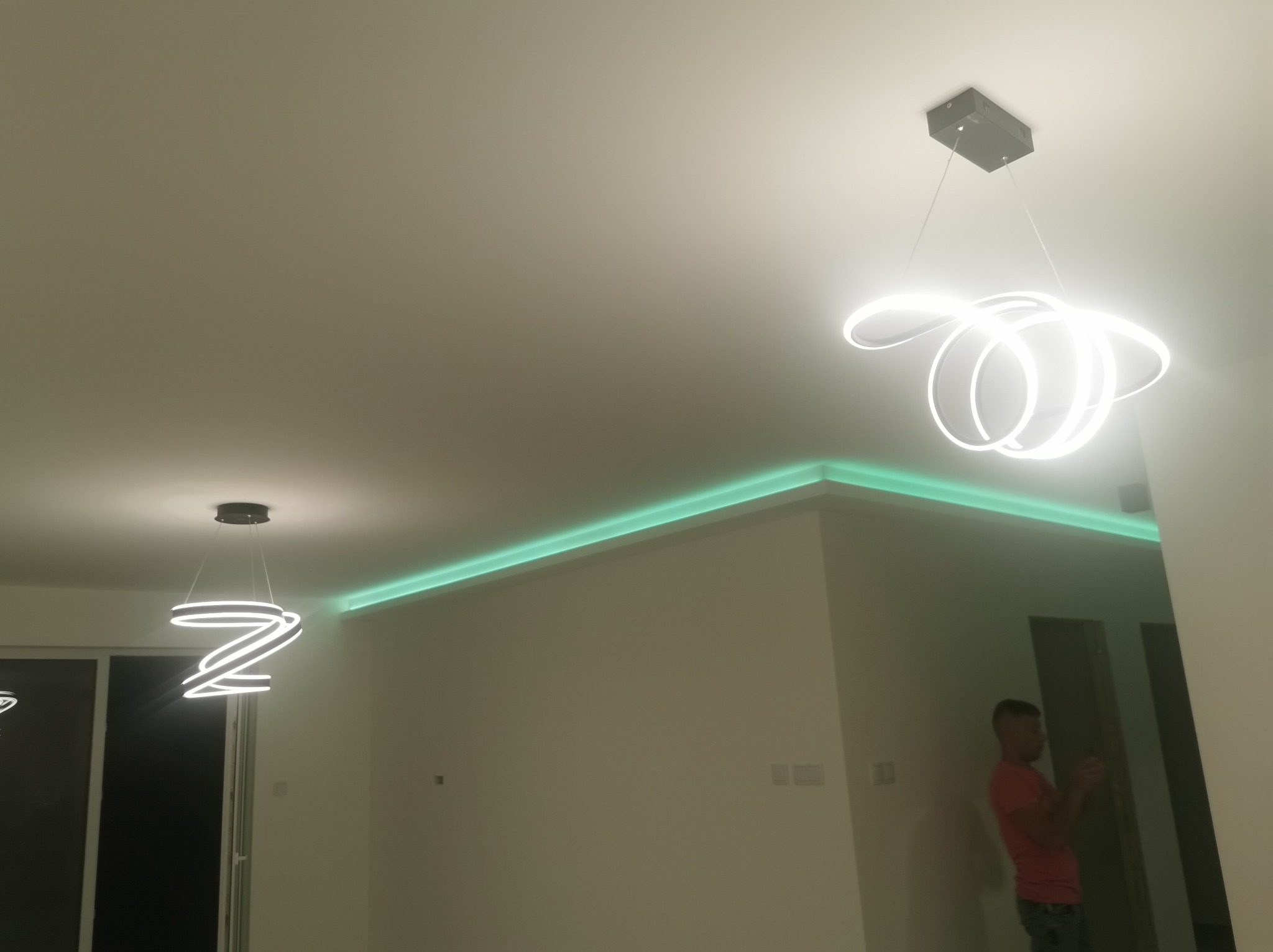 Nowoczesne oświetlenie wnętrza: Dwa wiszące, białe, spiralne żyrandole LED oraz zielone oświetlenie LED podsufitowe, widoczny fragment ściany i sufitu, oraz osoba w tle.