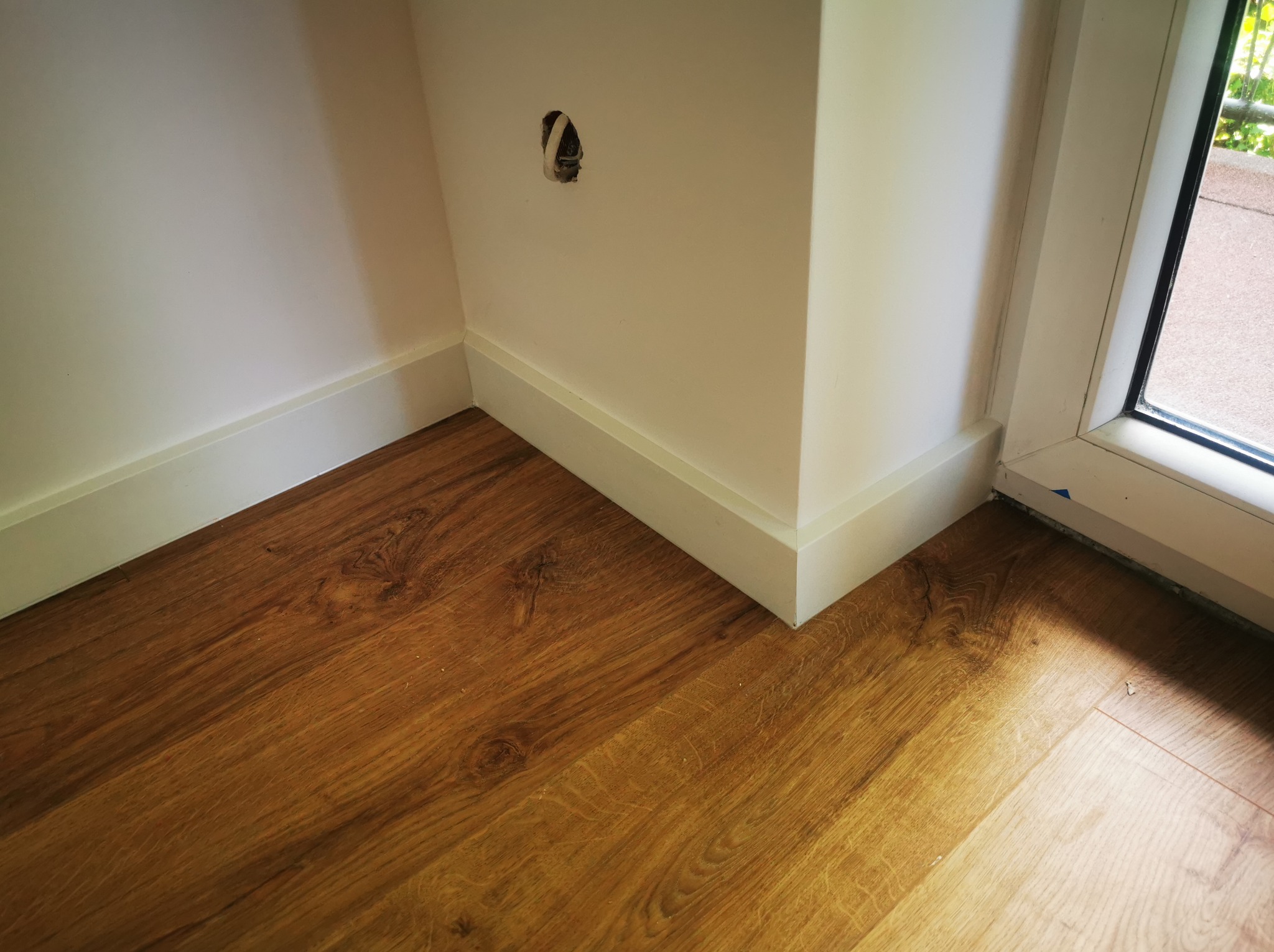 Fragment rogu pokoju z drewnianą podłogą, białą listwą przypodłogową i otworem na kabel w ścianie