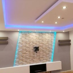 Prywatna - Nowoczesny salon z podświetlaną ścianą z kamienia dekoracyjnego, minimalistyczną białą szafką RTV i półkami ściennymi, oświetlenie sufitowe LED, okno balkonowe.