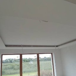 Prywatna - Wykończony sufit podwieszany z wnęką na oświetlenie, widoczne przewody elektryczne, ściany częściowo zagruntowane, duże okno z widokiem na zieleń.