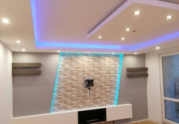 Nowoczesny salon z podświetlaną ścianą z kamienia dekoracyjnego, minimalistyczną białą szafką RTV i półkami ściennymi, oświetlenie sufitowe LED, okno balkonowe.