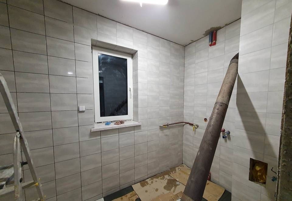 Wnętrze łazienki w trakcie wykańczania: jasne, kwadratowe płytki na ścianach, nowe okno PCV, wystające rury instalacyjne, zabezpieczona podłoga, widoczna drabina malarska.