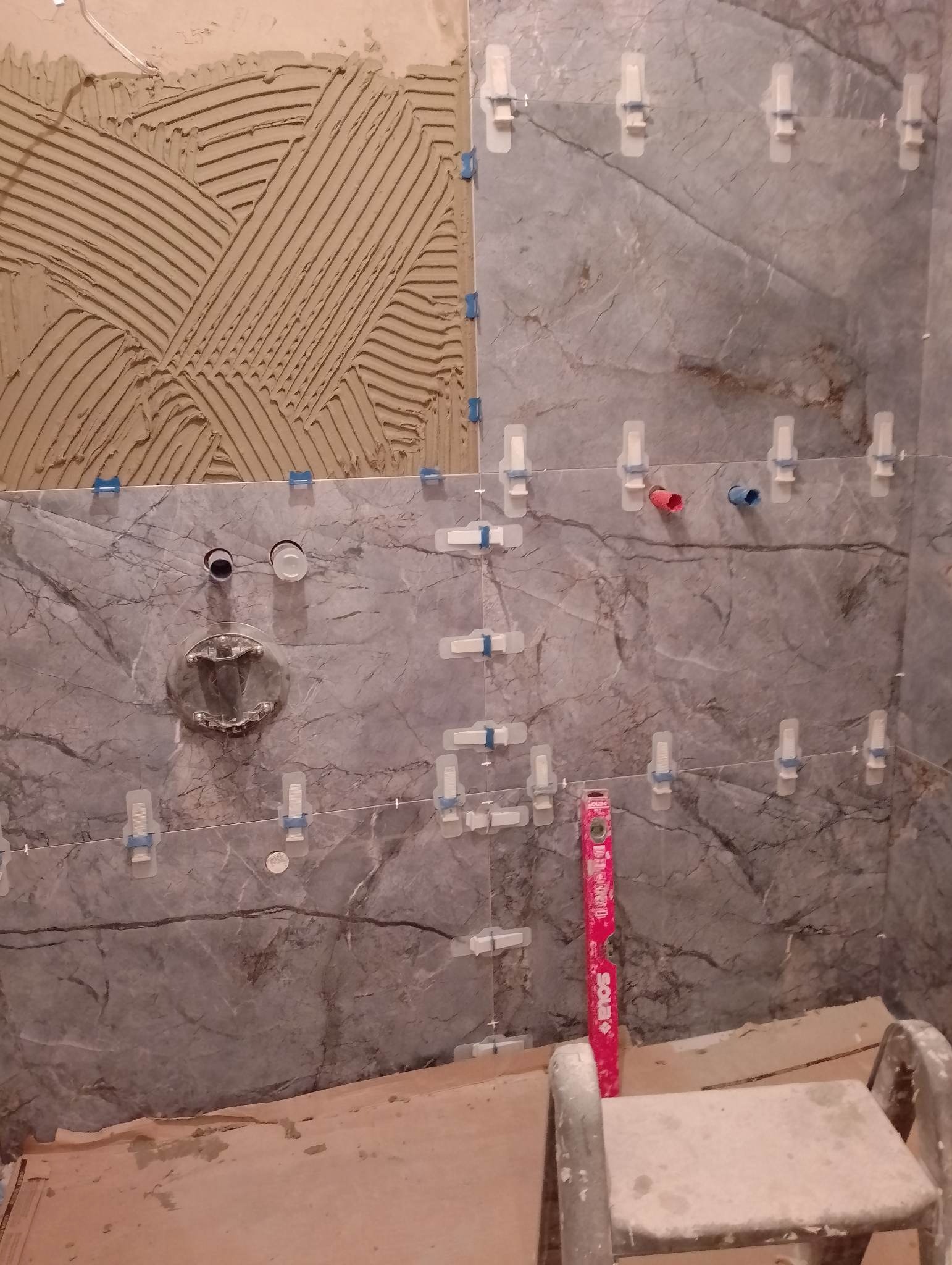 Montaż marmurowych płytek ściennych w łazience, widoczne klej, krzyżyki dystansowe, poziomica i przygotowane otwory na armaturę.