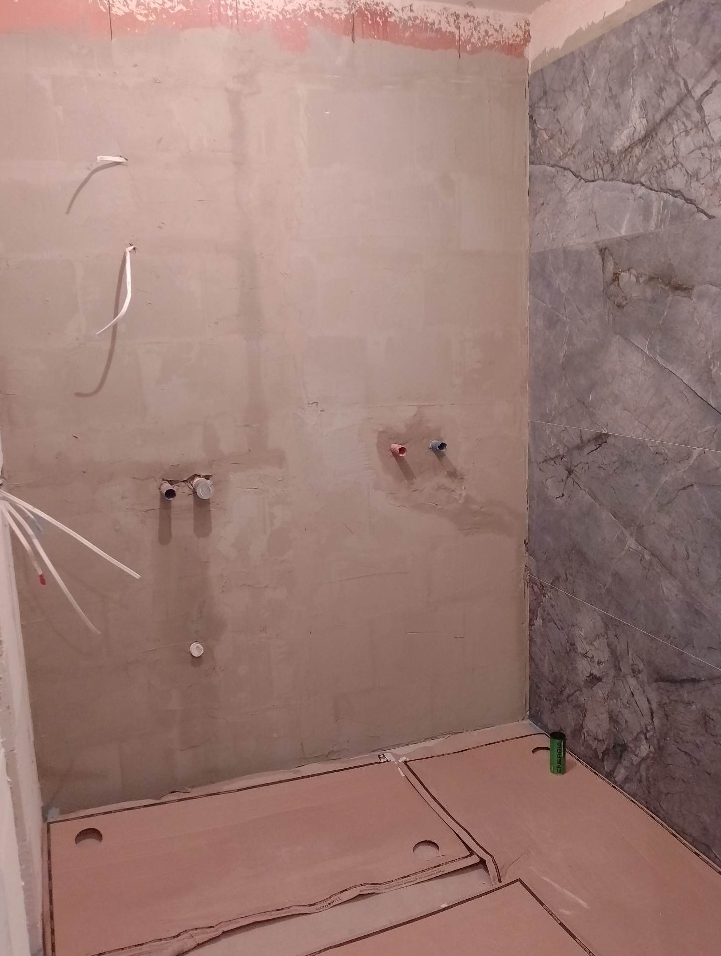 Niedokończona łazienka z szarą ścianą imitującą marmur, surową ścianą przygotowaną pod gładź, wystającymi rurami instalacji wodnej oraz rozłożonymi panelami pod ogrzewanie podłogowe. Widoczna...