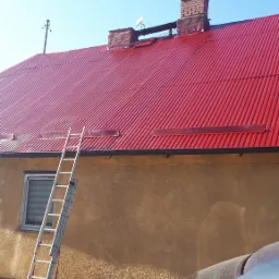 Czerwony dach z blachy falistej po renowacji, drabina oparta o ścianę budynku, komin z cegły w tle błękitne niebo.
