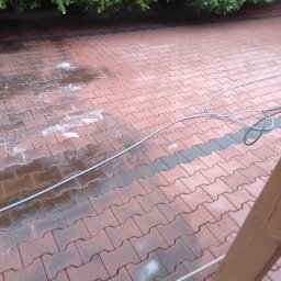 SOLARCELL - Częściowo umyta czerwona kostka brukowa z widocznymi śladami wody i piany, szary wąż ciśnieniowy oraz fragment drewnianej podpory w rogu kadru, w tle zielony żywopłot.