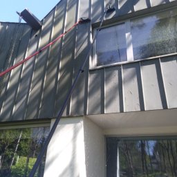 SOLARCELL - Długie lance z szczotkami czyszczą okna i metalowe panele elewacyjne nowoczesnego domu, woda spływa po szybach i elewacji.