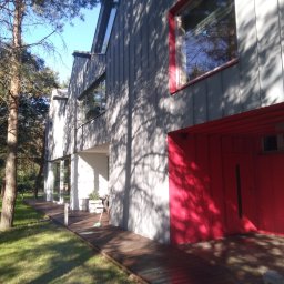 SOLARCELL - Nowoczesny dom z szarą elewacją i czerwonymi akcentami, drewnianym tarasem i widocznym cieniem drzew na ścianie.