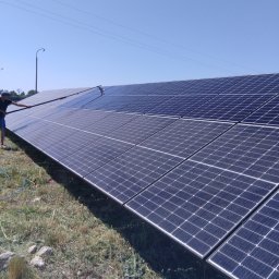 SOLARCELL - Czyszczenie paneli słonecznych z użyciem szczotki teleskopowej i węża doprowadzającego wodę, widoczne trawy i niebo.