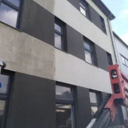 Mycie elewacji budynku z wyraźnym kontrastem czystej i brudnej powierzchni, widoczny podnośnik koszowy.