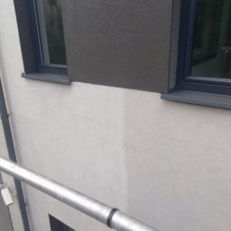 SOLARCELL - Fragment elewacji budynku z widocznym efektem czyszczenia, gdzie górna część jest szara i brudna, a dolna biała i czysta, z oknami w ciemnych ramach.