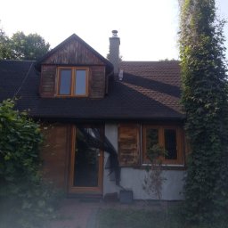 SOLARCELL - Dom jednorodzinny z ciemnym dachem i drewnianą elewacją w trakcie przygotowań do renowacji, z widocznymi osłonami na oknach i drzwiach, otoczony bujną roślinnością.