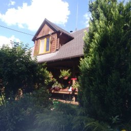 SOLARCELL - Dwupiętrowy dom z brązową dachówką i drewnianą elewacją, widoczny zza gęstej zieleni krzewów i drzew, z oknem na poddaszu z żółtą ramą i kwiatami w wiszących doniczkach na werandzie.