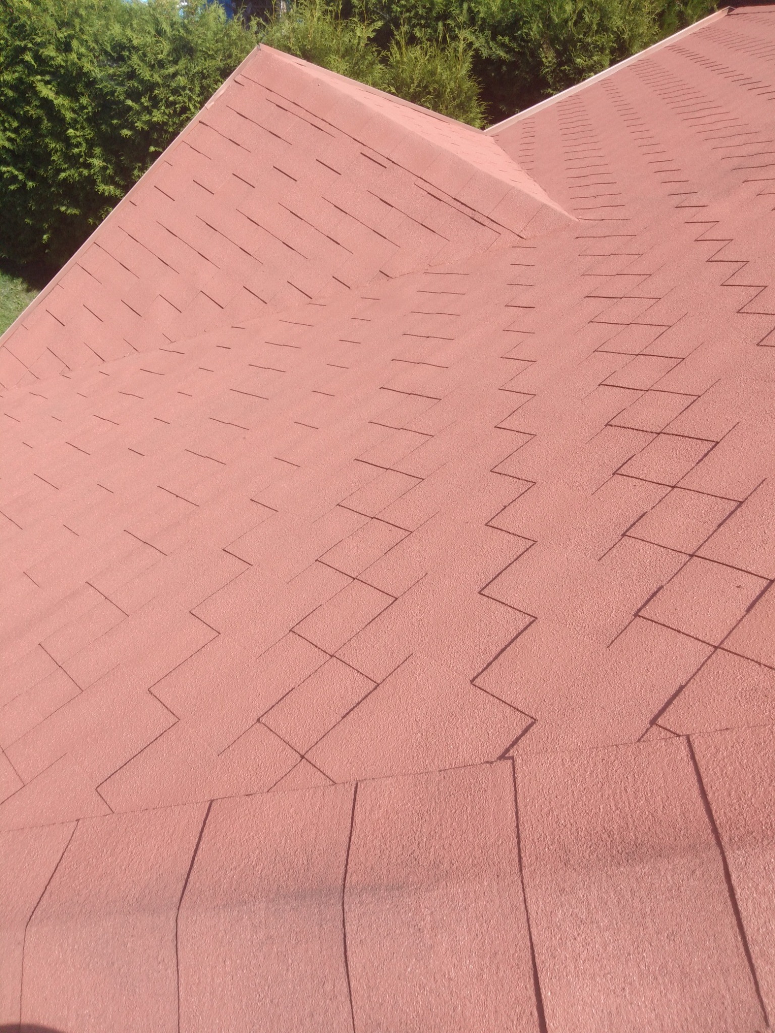 Czerwony dach pokryty gontem bitumicznym, widok z góry, zarys zieleni drzew w tle.