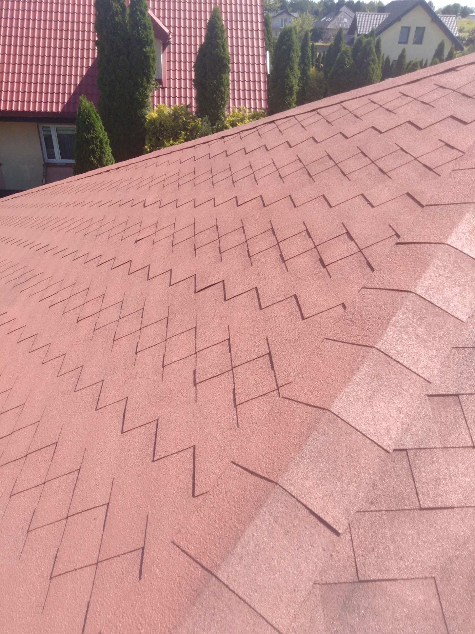 Czerwony dach pokryty gontem bitumicznym w zygzak, widok z bliska, w tle zabudowania i drzewa.