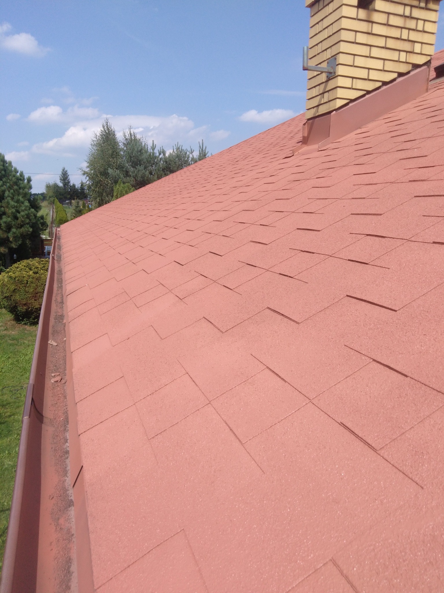 Czerwony dach pokryty gontem bitumicznym, widoczny ceglany komin i rynna z zabrudzeniami, błękitne niebo w tle.