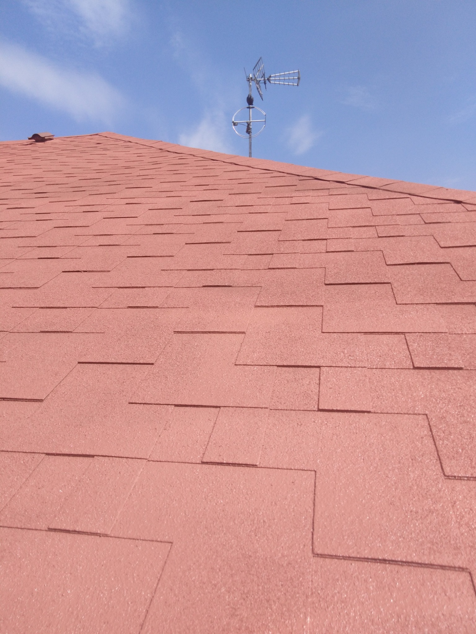 Czerwony dach pokryty gontem bitumicznym, z widoczną anteną telewizyjną na szczycie, na tle błękitnego nieba z delikatnymi chmurami.