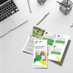 Rozłożona, trójdzielna ulotka reklamowa na białym biurku obok laptopa, długopisów i okularów, prezentująca ofertę firmy PMP dla młodych przedsiębiorstw, z zielono-żółtą grafiką.